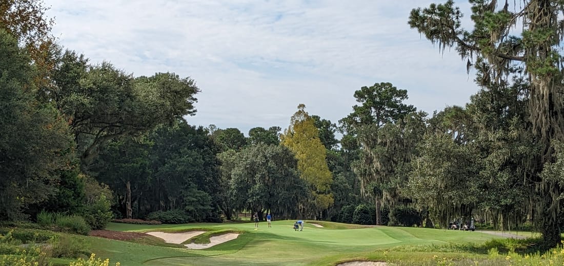 Live oaks draped in Spanish moss framing a narrow fairway corridor at Caledonia Golf and Fish Club in Pawleys Island, South Carolina, with the clubhouse visible beyond the closing green.