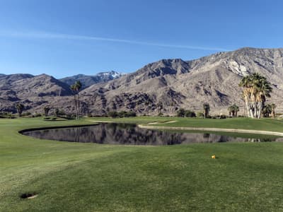 A desert golf course fairway in the Coachella Valley at dusk, with bare rocky mountains behind the green and long shadows stretching across the turf.
