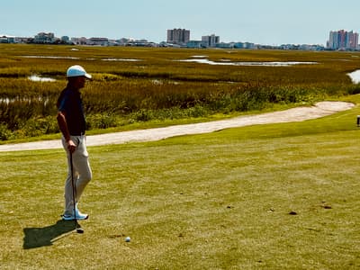 Elevated tee at Tidewater Golf Club looking out over Cherry Grove salt marsh toward the Atlantic Ocean, with tidal creeks winding through spartina grass below the fairway.
