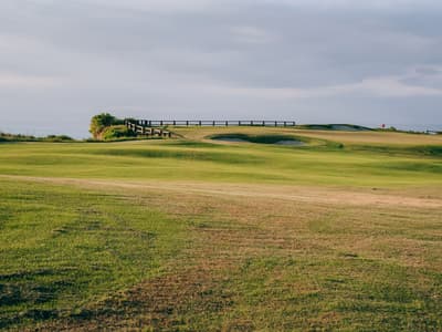 A long par 3 green set at a diagonal to the tee, canting away from front-right to back-left, guarded by a deep bunker cut into the front-left face of the plateau.